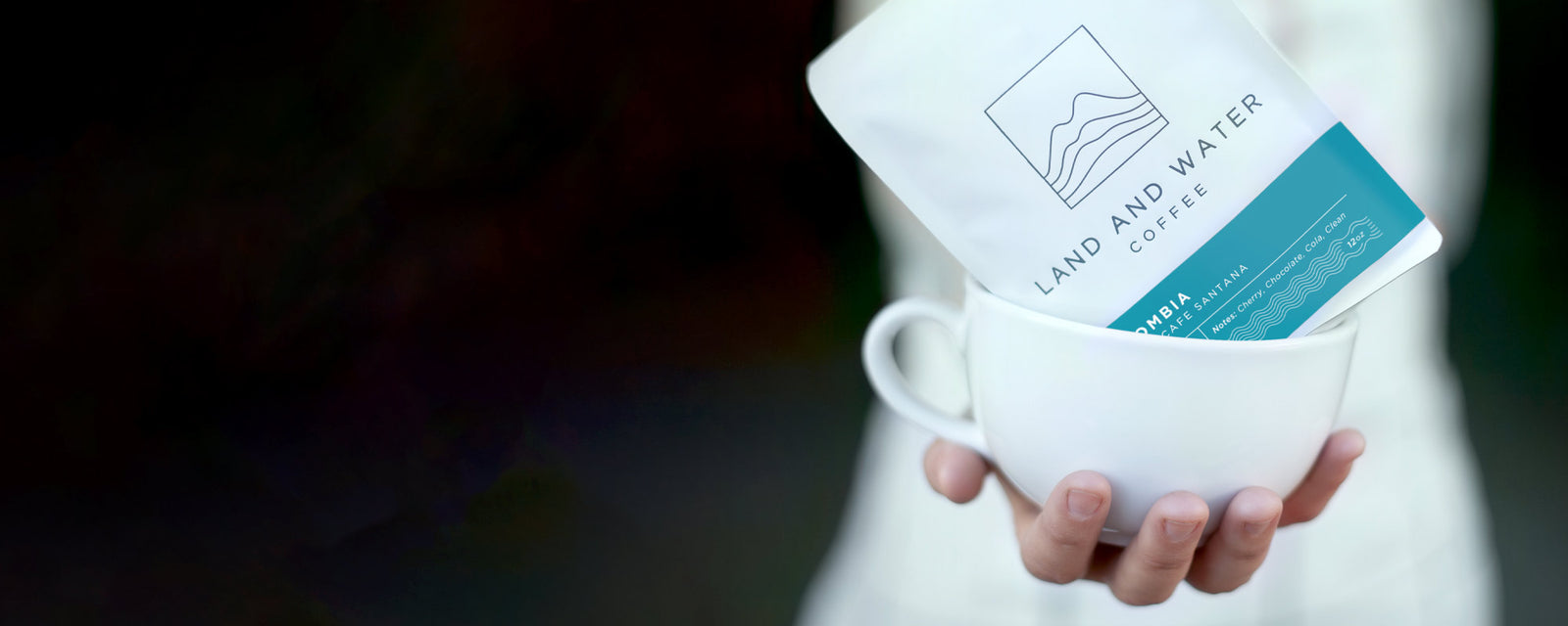 a closeup of a barista's hand holding a white ceramic latte cup with an entire bag of whole bean coffee inside the cup instead of brewed coffee. Coffee bag is white with dark logo and a blue Colombia origin label. The foreground is in focus and the barista's body is blurred in the background. 