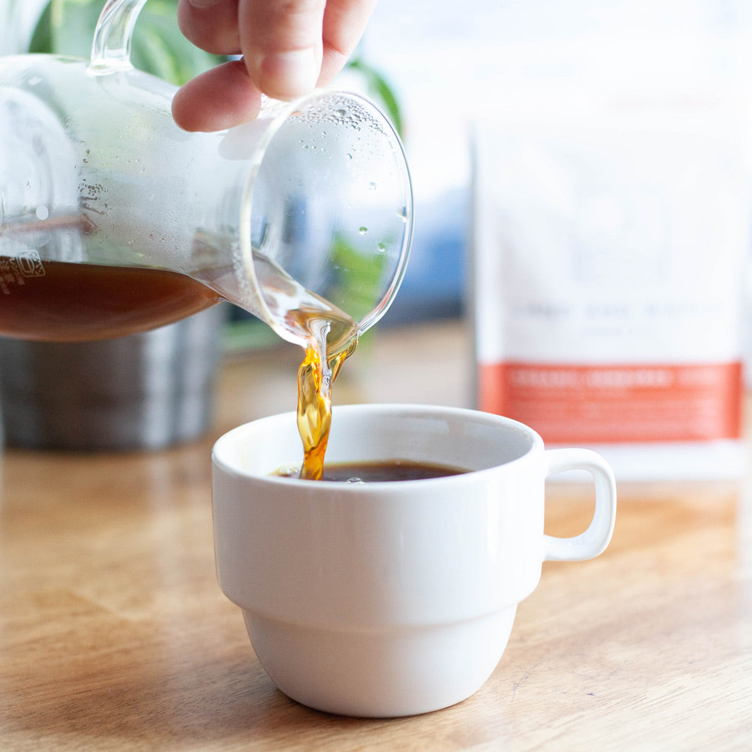 a hand pouring a brewed coffee from a clear glass carafe into a small white ceramic mug on top of a blonde wooden tabletop; the coffee is light in color and the carafe is steaming from the heat; there's a blurred plant next to a white coffee bag with a red label in the background.