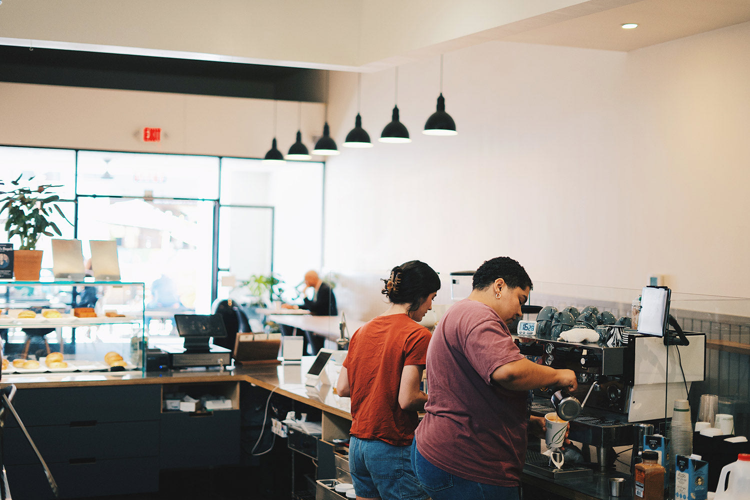 Inside our downtown cafe with two baristas working behind the espresso bar; white walls with hanging black pendant lights. 